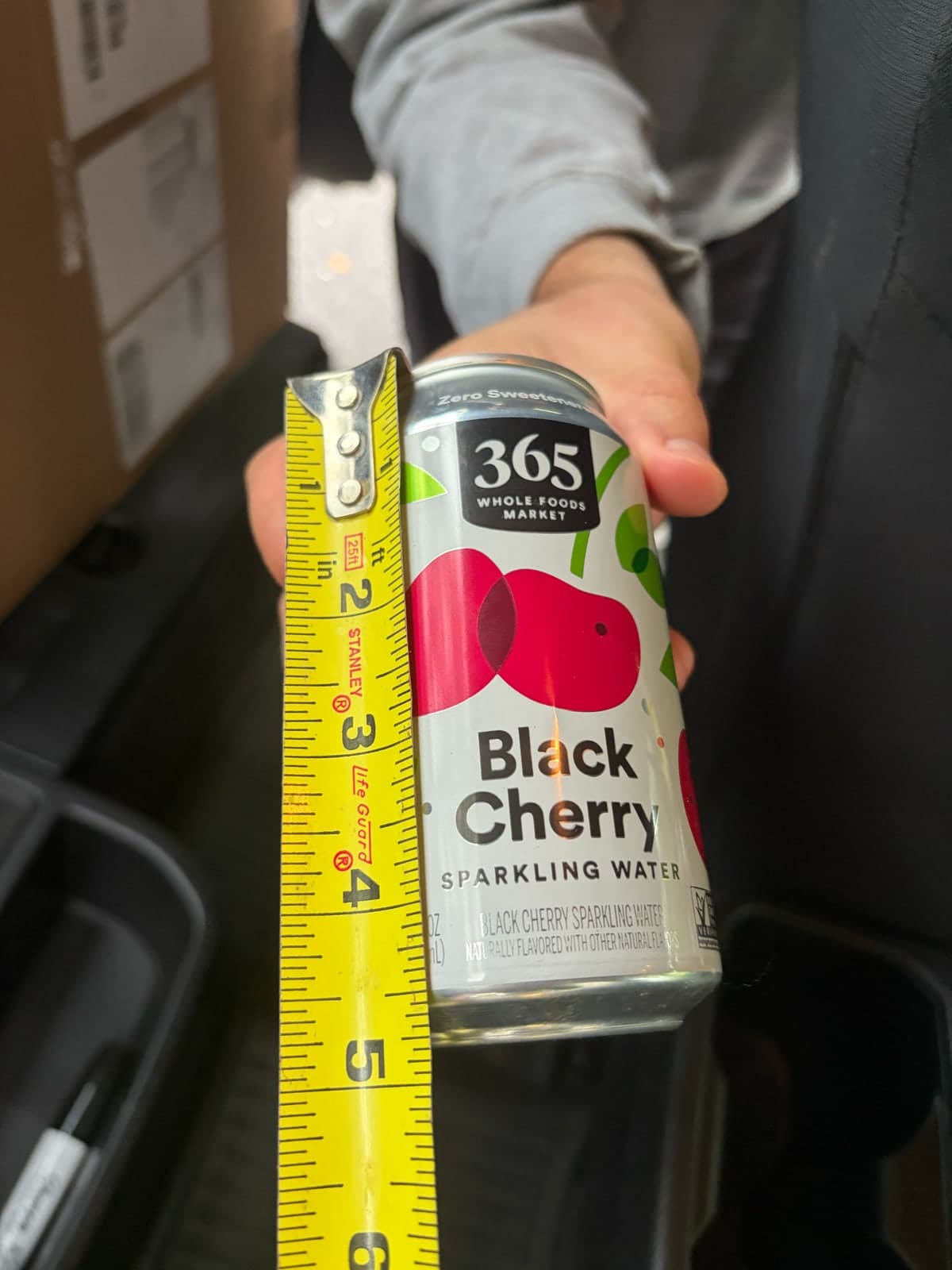 A hand holds a drink can beside a yellow tape measure as a size reference.