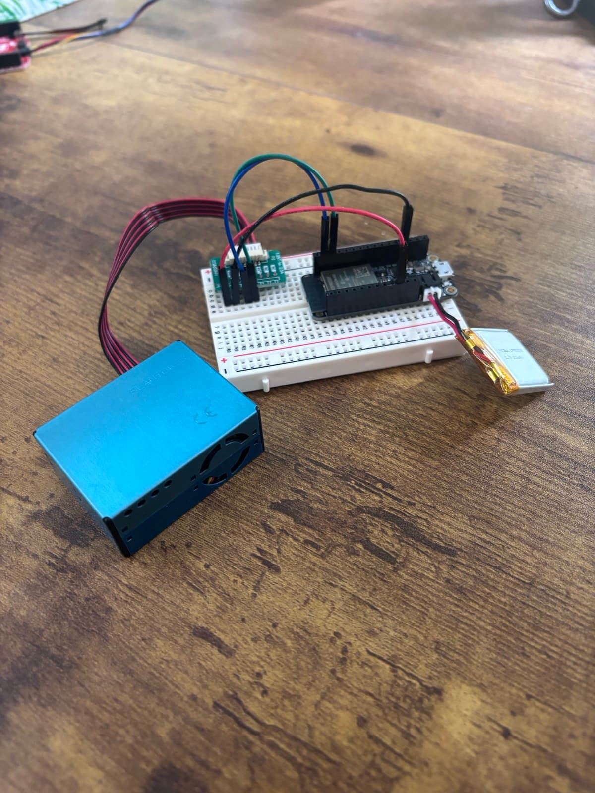 An Adafruit Feather on a breadboard, wired with jumper wires to a Plantower particulate sensor, powered by a small gold LiPo battery, on a wooden desk.